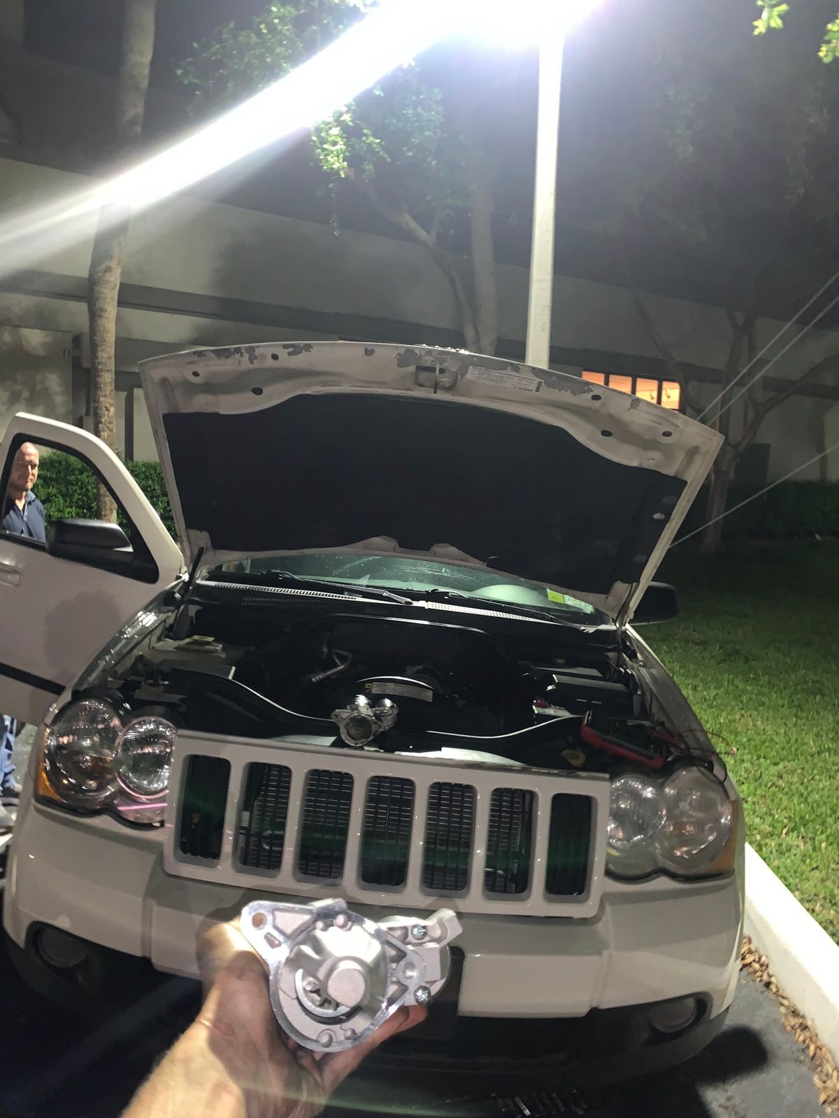 Technician holding a starter motor during a night repair on a Jeep Grand Cherokee